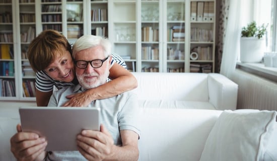 woman hugging man from behind couch who is looking at a tablet computer