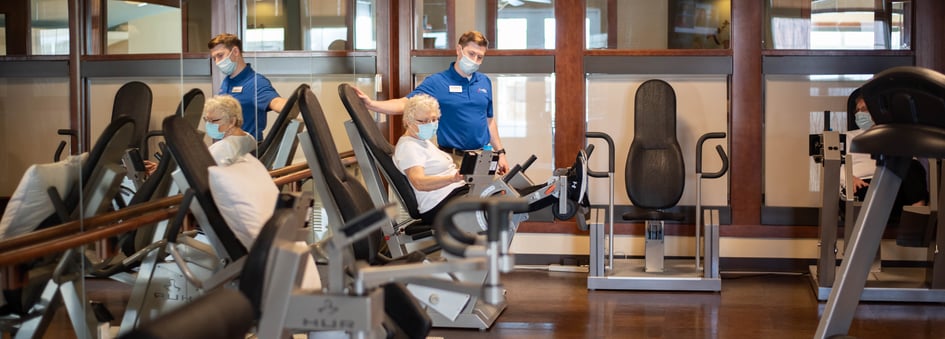 Senior woman sits on workout equipment next to staff member